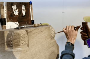 A person is using a chisel and hammer to carve intricate floral designs into a natural stone surface. A reference sketch of flowers is clipped nearby, adding to the timeless beauty of this focused craftsmanship in stone carving.
